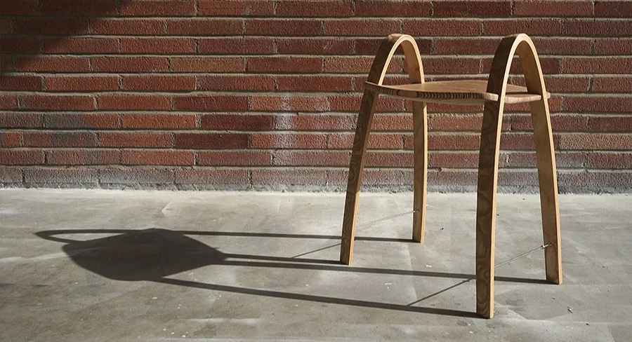 A low wooden chair with two tall bent-plywood bows for legs, casting a shadow on a brick wall
