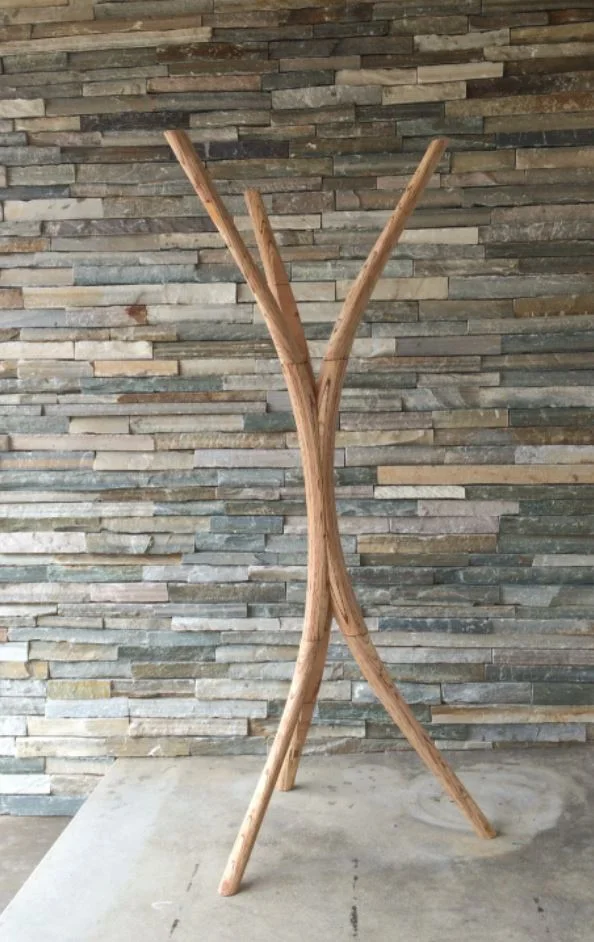 A tall wooden coat rack of slender curved limbs crossing at the middle, against a stone wall