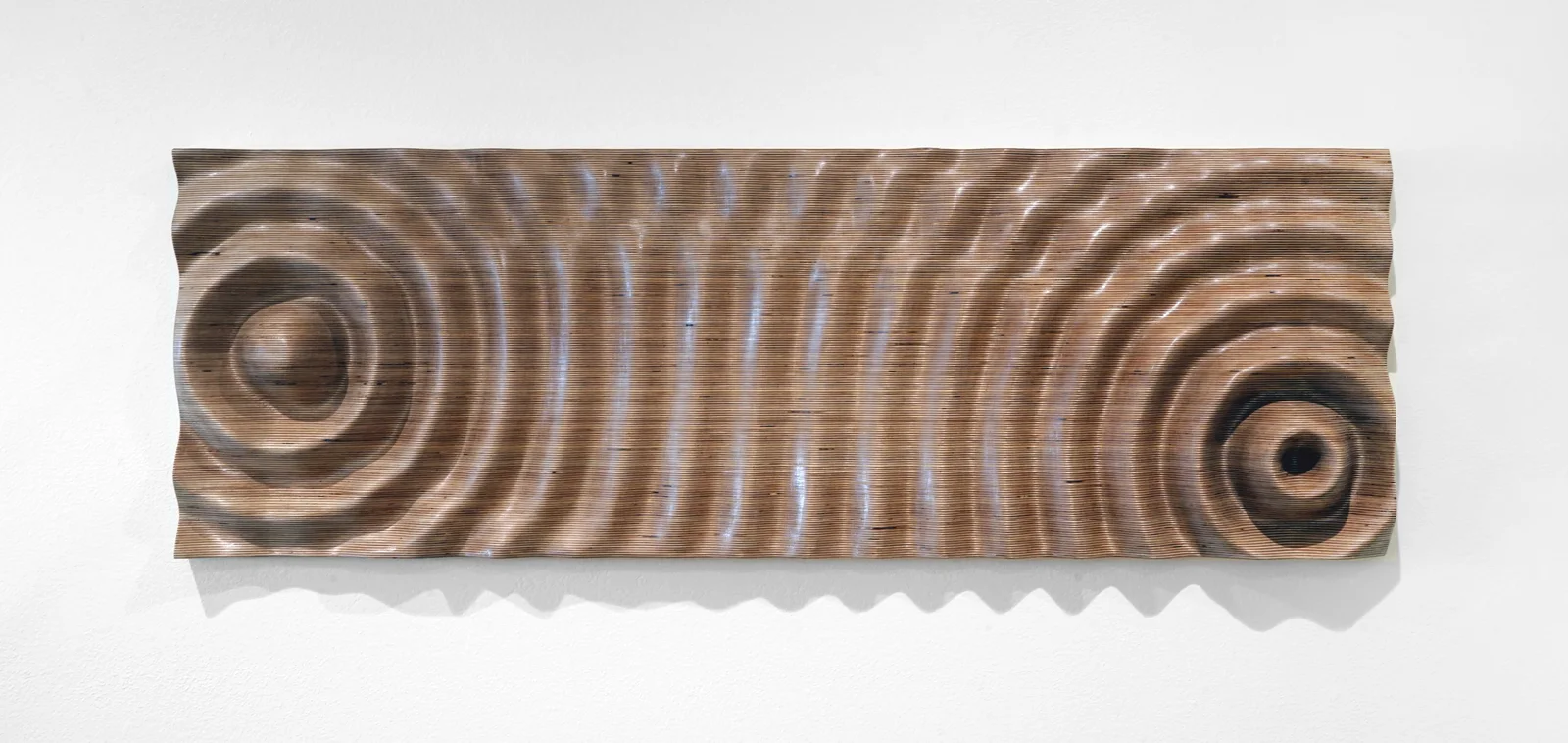 A long wooden panel milled into concentric ripples spreading from two points, its laminated layers showing as rings