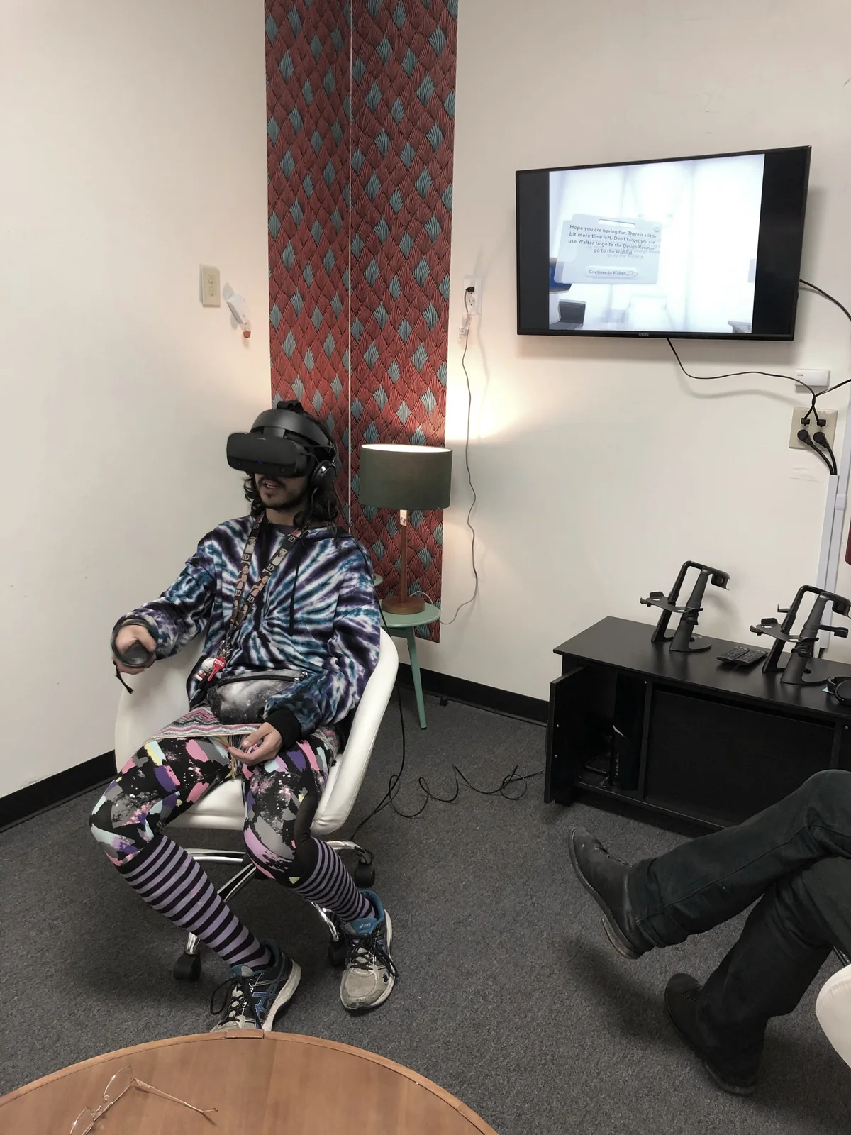 A participant in a headset with a controller, the Design Room on the screen beside them