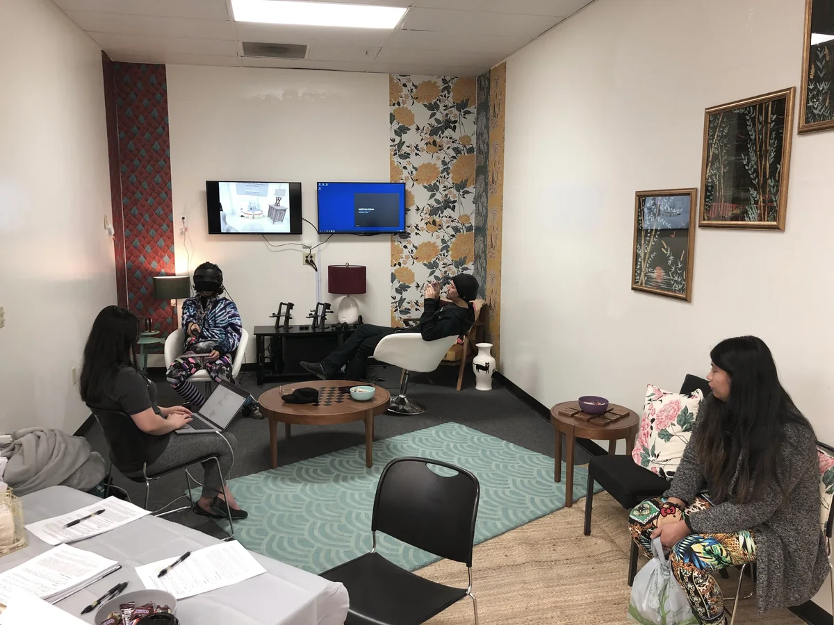Two participants in VR headsets at a table, their view shown on two screens on the wall
